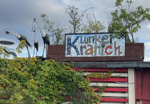 Auf dem Foto ist ein kleines Gebäude mit einem Schild. Darauf steht Klunkerkranich. Links daneben stehen 3 Kranichfiguren. Über dem Schild wachsen Sträucher. Foto: Katzenauge