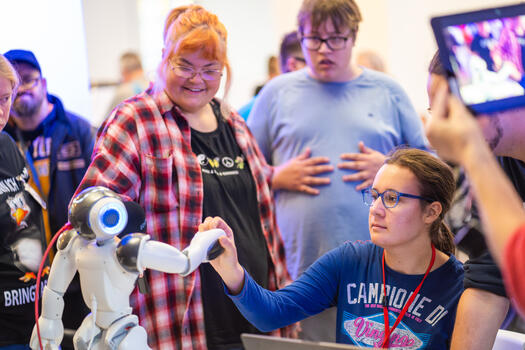 Auf einer Veranstaltung mit vielen Menschen: Eine junge Frau schüttelt einem Roboter die Hand. Foto: Dennis Lenz