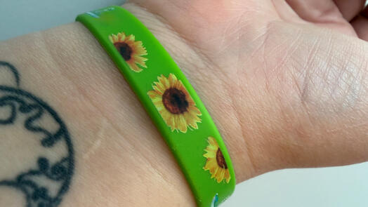 Auf dem Bild sieht man einen Ausschnitt von einem Arm. Der Arm trägt ein grünes Armband mit Sonnenblumen.