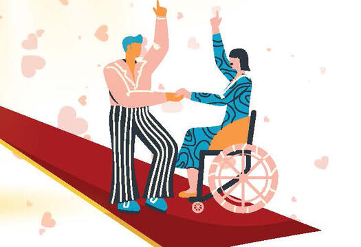Auf dem Bild sind ein Mann und eine Frau zusammen auf einem roten Teppich. Die Frau sitzt im Rollstuhl. Der Mann steht vor ihr. Sie halten sich an der Hand. Die andere Hand zeigt mit dem Zeigefinger nach oben.