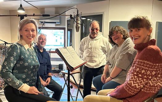 5 Menschen sitzen in einem Tonstudio um ein Mikrofon herum.