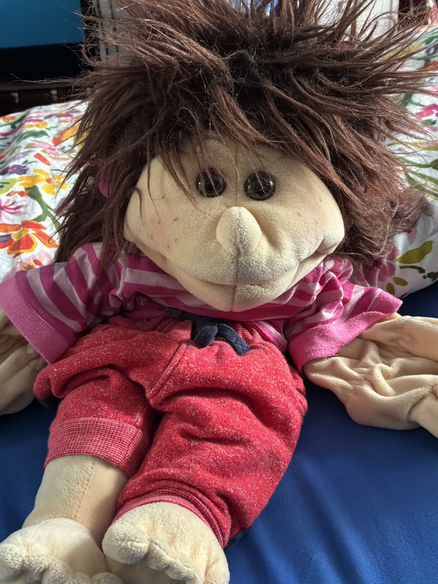 Eine Bauchredner-Puppe: Sie hat dunkle Haare, ein rosa Ringel-T-Shirt, eine rote Hose und sitzt auf einem Bett.