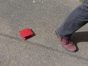 Eine rote Brieftasche liegt auf dem Boden. Davor ist ein weggehendes Bein.