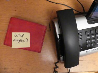 Eine Brieftasche liegt auf dem Tisch mit einem Zettel. Daraus steht: Wird abgeholt. Daneben steht ein schwarzes Telefon.