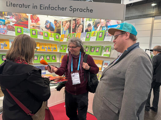 Ein Stand mit Literatur in Einfacher Sprache auf der Leipziger Buchmesse 2025. 3 Menschen stehen vor bunten Büchern. Ein Reporte rmacht ein Interview.