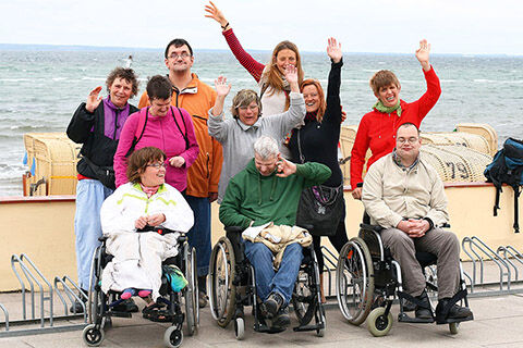 Eine Gruppe von Urlaubern, darunter drei Rollstuhlfahrer, winken in die Kamera - im Hintergrund Meer und Strandkörbe